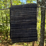 A dark woven blanket with horizontal stripes of black, navy, and subtle tan flecks, hanging from a tree branch in a forest setting.