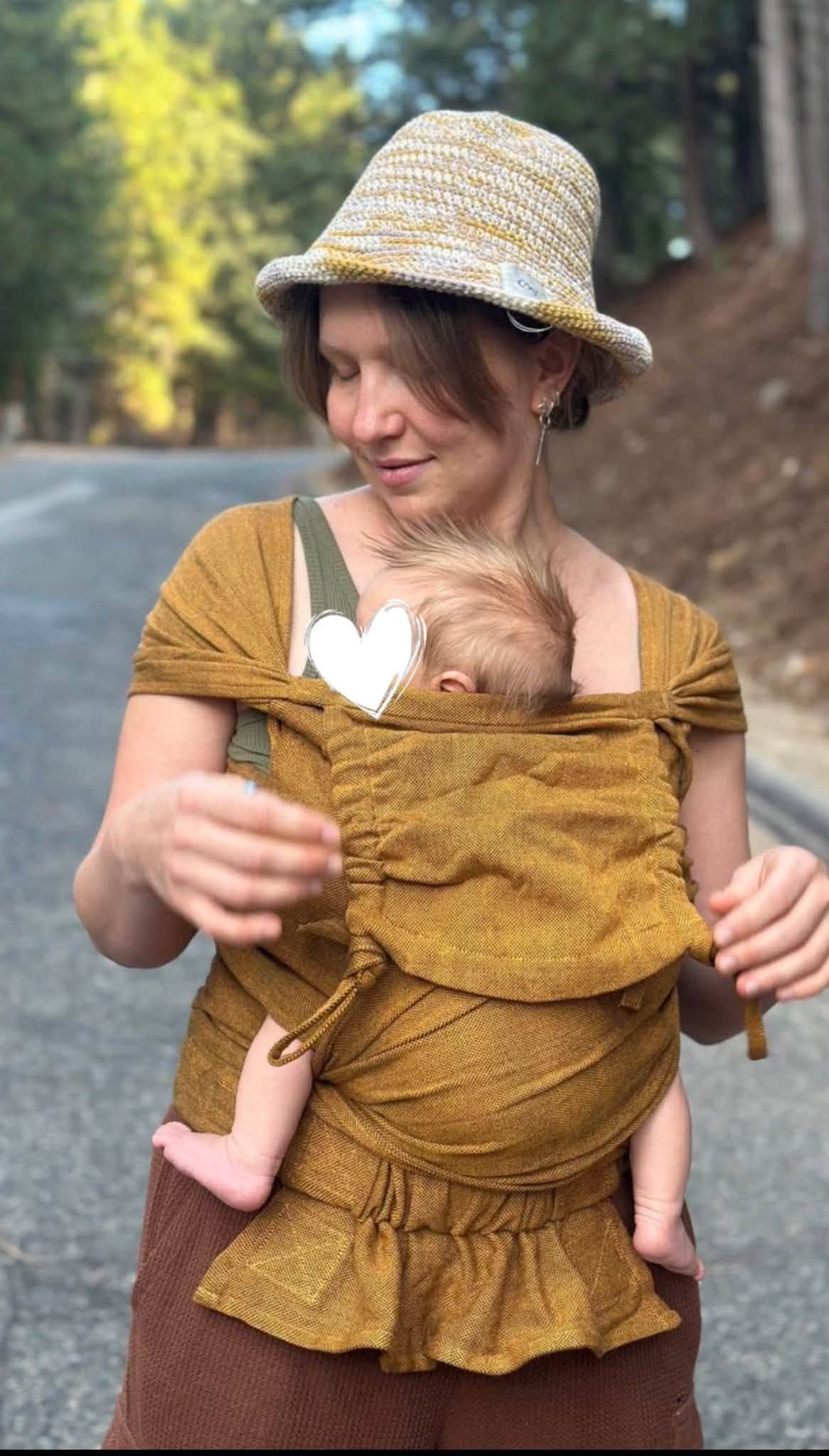 A woman wearing a straw hat and a mustard yellow baby wrap carrier with a baby inside, standing outdoors on a road with trees in the background.