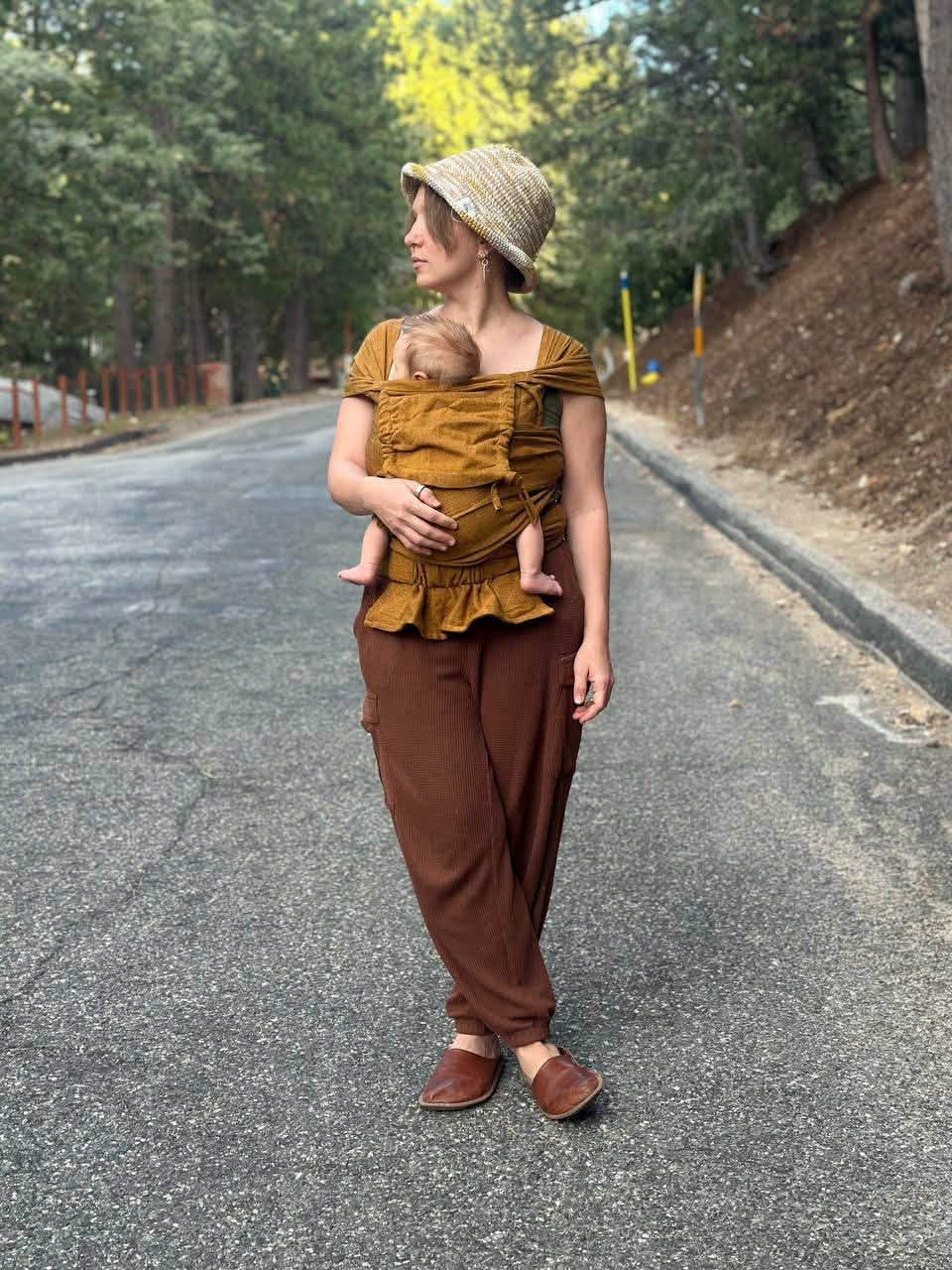 A woman wearing a mustard yellow brown baby wrap carrier  and brown pants stands on an asphalt road surrounded by trees, holding a baby in a woven straw hat and baby carrier outfit.