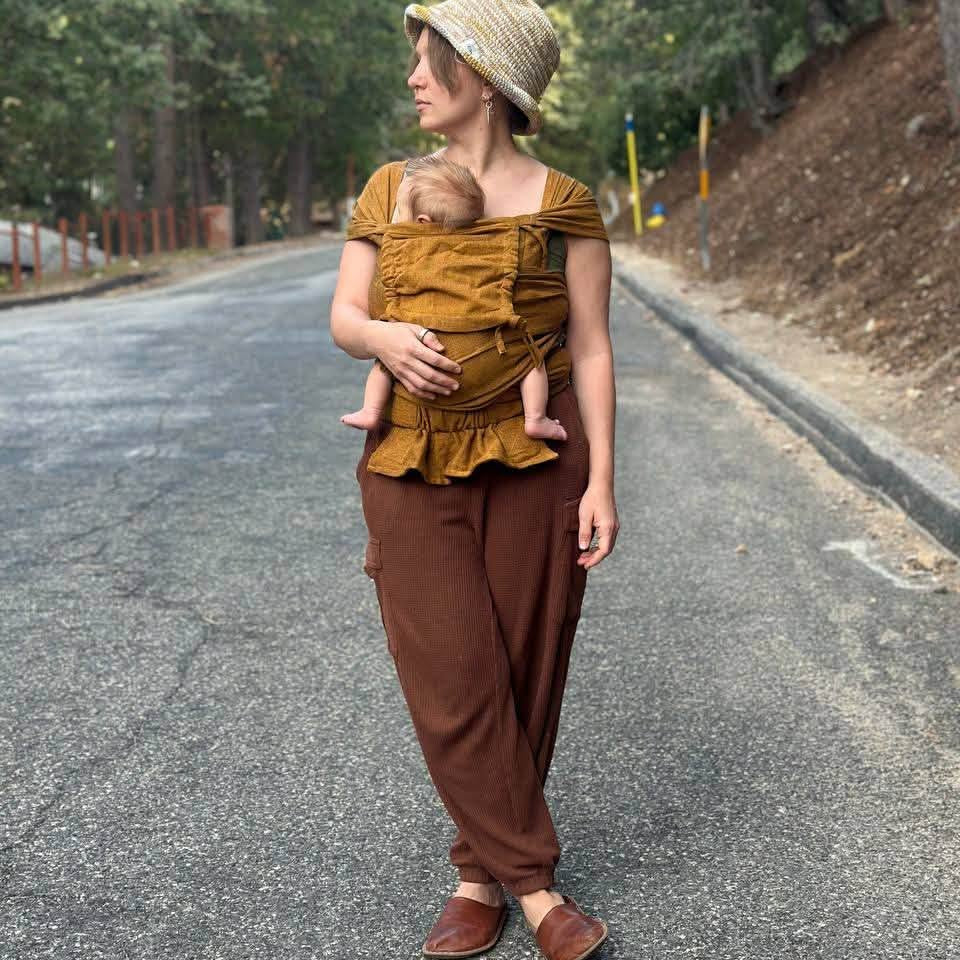 A woman wearing a mustard yellow brown baby wrap carrier  and brown pants stands on an asphalt road surrounded by trees, holding a baby in a woven straw hat and baby carrier outfit.