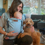 Woman holding a baby in a carrier and a dog in a living room.