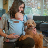 Woman holding a baby in a carrier and a dog in a living room.
