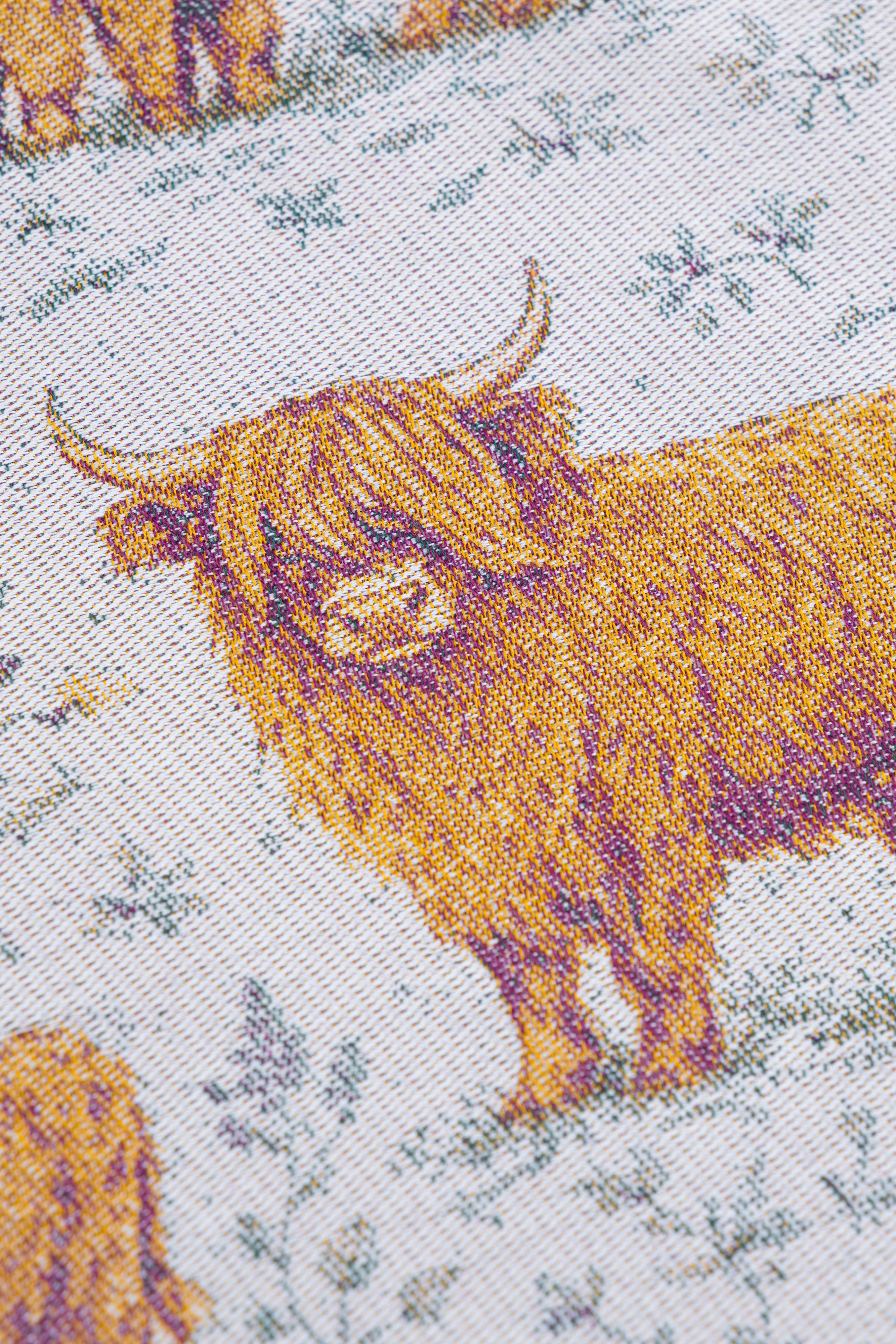 Close-up of a woven textile featuring Highland cows in golden yellow and brown tones on a light off-white background with muted blue-gray floral details.